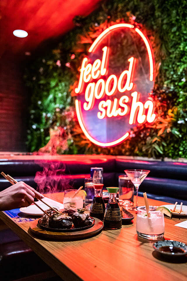 Sizzling River Rock Beef on a table with cocktails in front of a neon Feel Good Sushi sign.